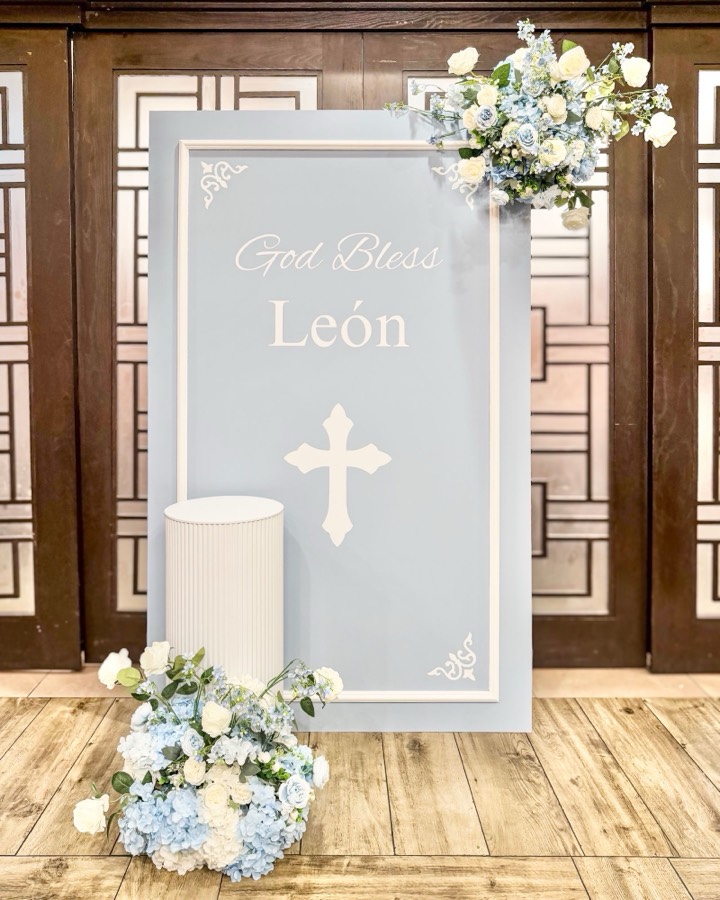 Powder-blue framed panel reading “God Bless León” with a white cross, an ivory fluted pedestal and cascading blue hydrangea and white rose arrangements.