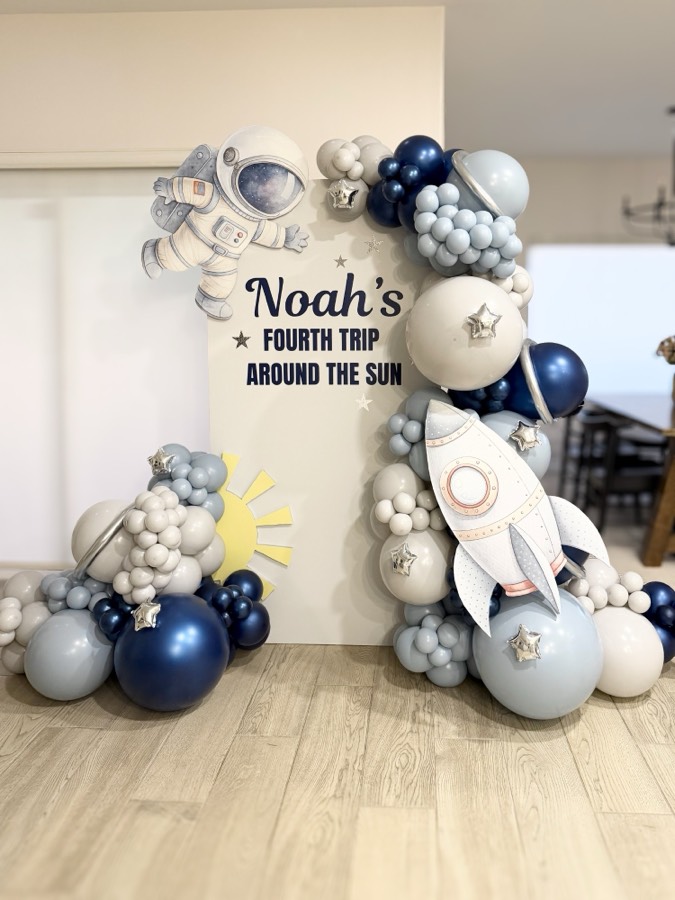 Ivory panel reading “Noah's Fourth Trip Around the Sun” with an illustrated astronaut and rocket, surrounded by navy, pale blue and grey balloons with silver star accents.