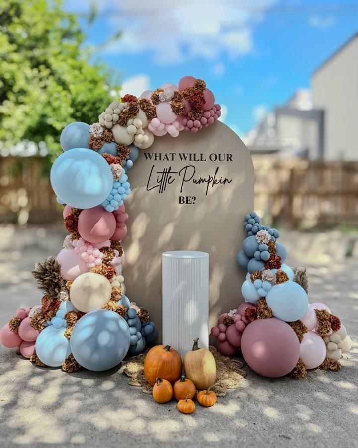 Taupe arch backdrop reading “What will our little pumpkin be?” framed by dusty blue, rose and cream balloons with dried florals and pumpkins at the base.