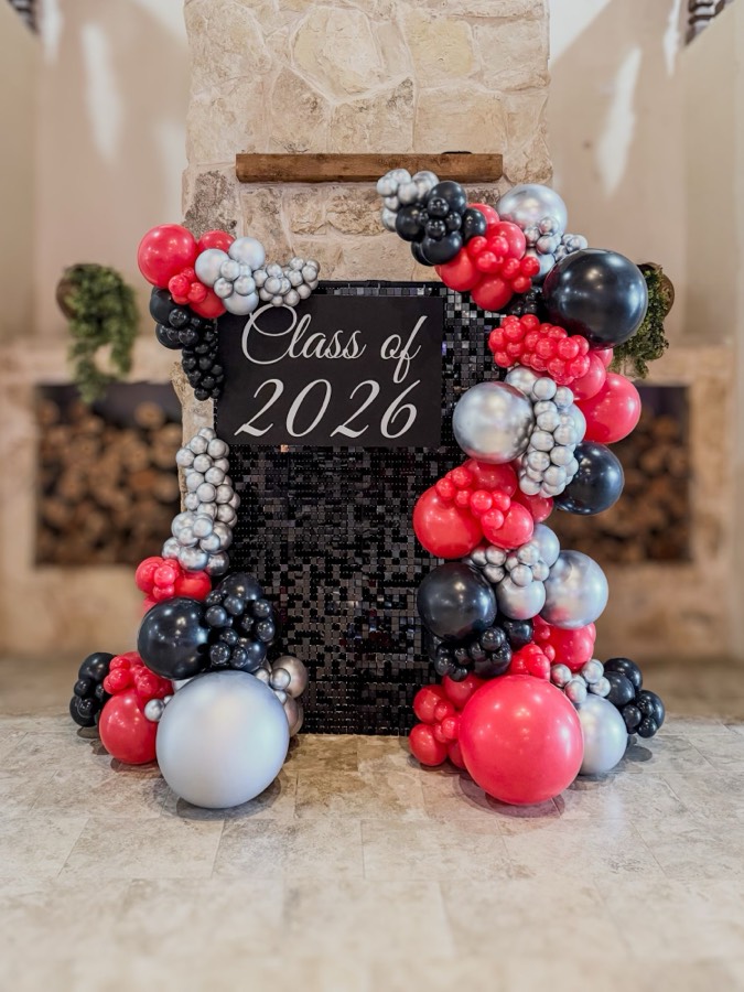 Black sequin shimmer wall with a “Class of 2026” sign, framed by black, red and chrome silver balloon garlands.
