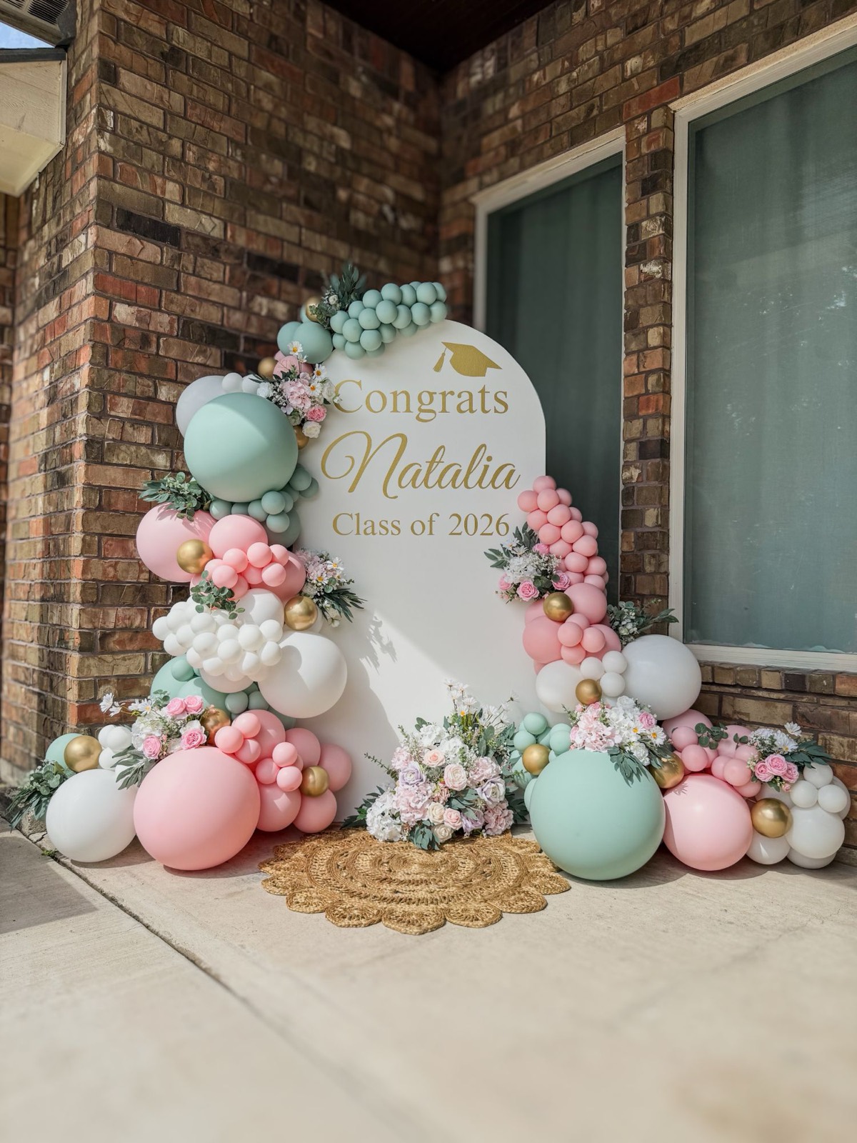 Ivory arch backdrop reading “Congrats Natalia, Class of 2026” in gold, framed by sage, blush, white and gold balloon garlands with pink and white florals.