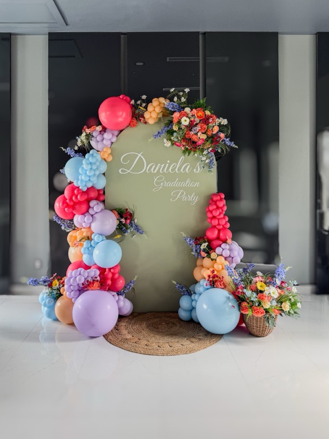 Sage arch backdrop reading “Daniela's Graduation Party” with coral, lavender, peach and sky-blue balloon garlands woven with wildflowers beside a flower basket.
